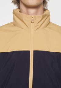 Person som bär en jacka med hög krage och dragkedja, med en tan-färgad överdel och mörk marinblå underdel, avklippt vid halsen och axlarna.