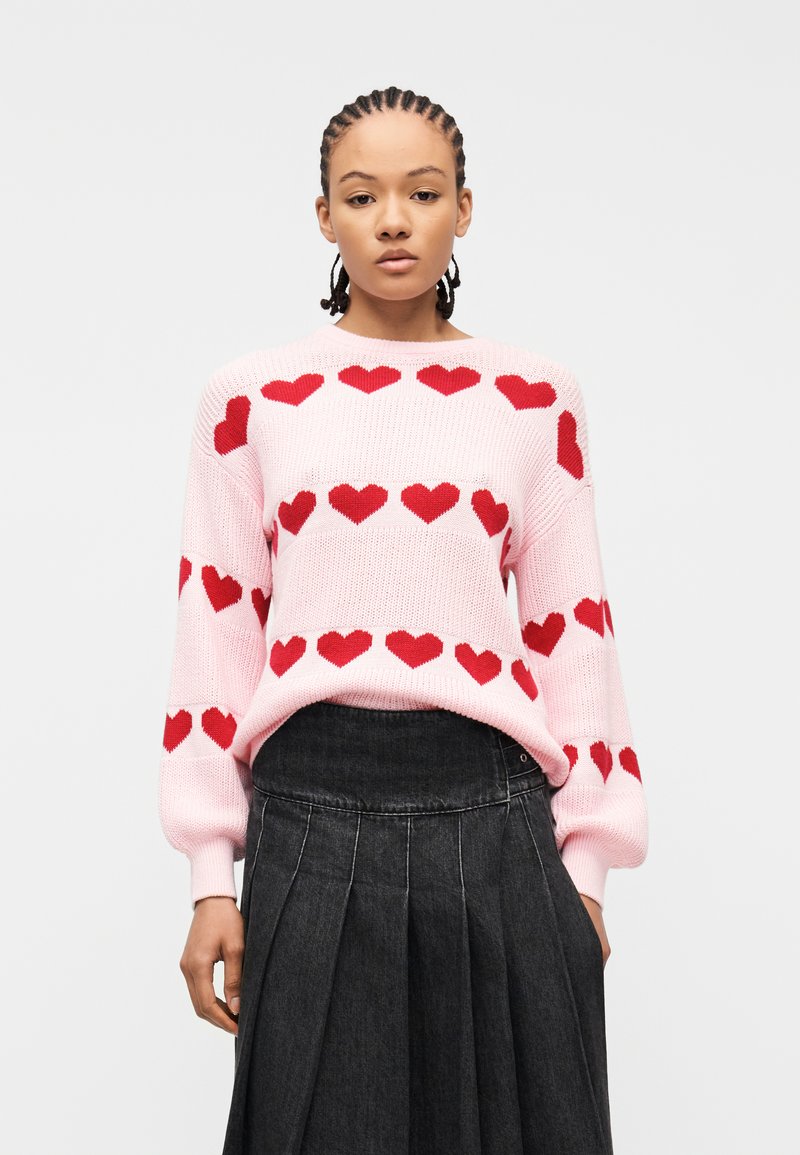 Νεαρό άτομο με πλεξούδες φοράει ροζ πουλόβερ με κόκκινη καρδιά και μαύρη φούστα με πιέτες και ψηλή μέση, πάνω σε λευκό φόντο.