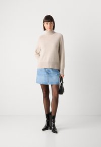 Beige coltrui met geribde zoom; lichtblauwe denim mini-jurk; zwarte enkellaarsjes; houdt een kleine zwarte handtas vast.