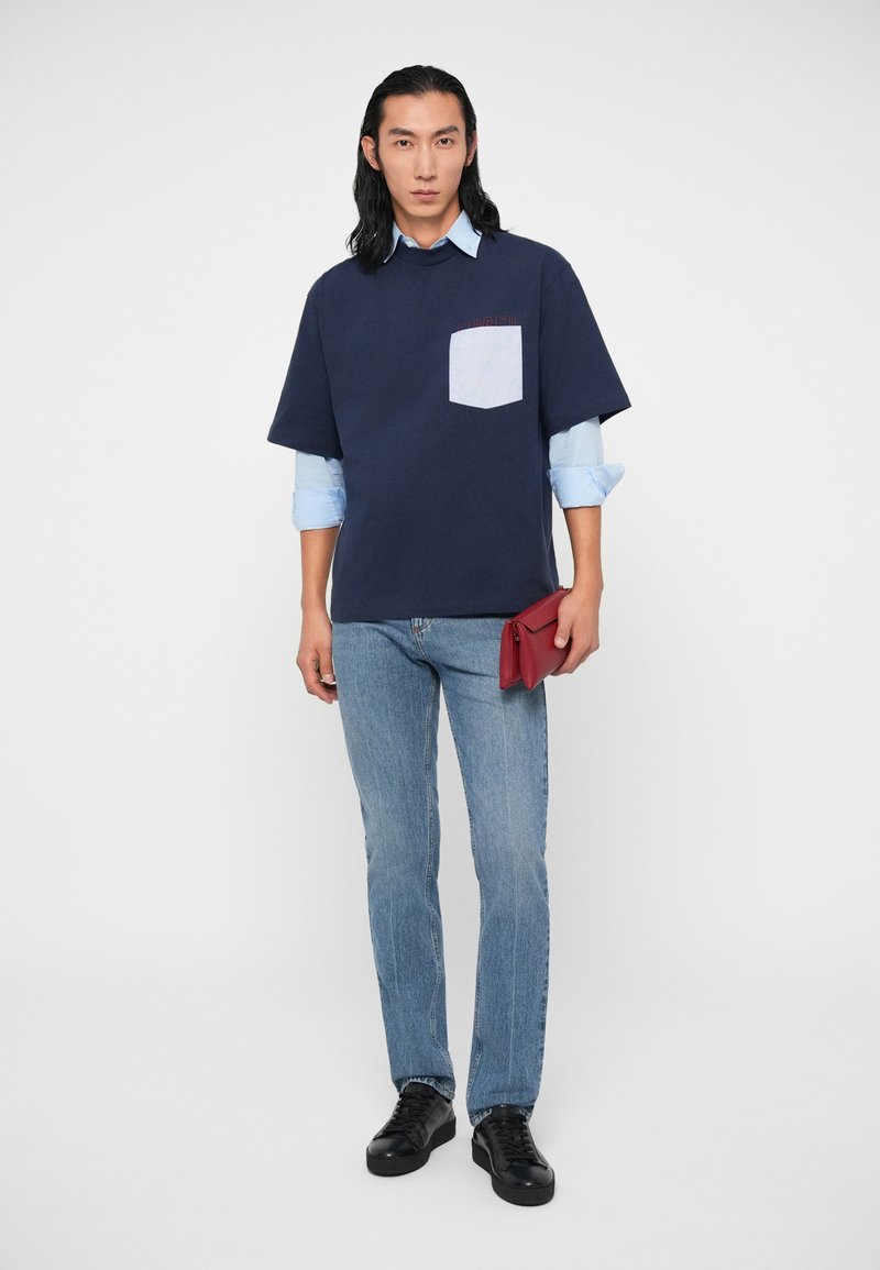 Mann mit langen schwarzen Haaren, der ein übergroßes navyblaues T-Shirt über einem hellblauen Hemd trägt, helle Jeans und schwarze Turnschuhe trägt und eine rote Clutch in der Hand hält.