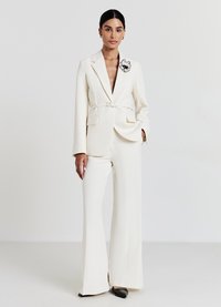 Completo bianco su misura con vita cintata, pantaloni a gamba larga e scollatura a V profonda. Presenta un accento di spilla floreale nera sul risvolto.