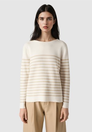 Vrouw met lang donker haar die een wit trui met lange mouwen draagt met beige horizontale strepen en beige broek, staand tegen een grijze achtergrond.