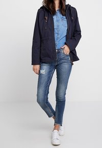 Giacca a cappuccio blu navy con zip frontale, due tasche sul petto, indossata sopra una camicia di jeans azzurri chiari. Abbinata a jeans skinny strappati blu medio e sneakers bianche.