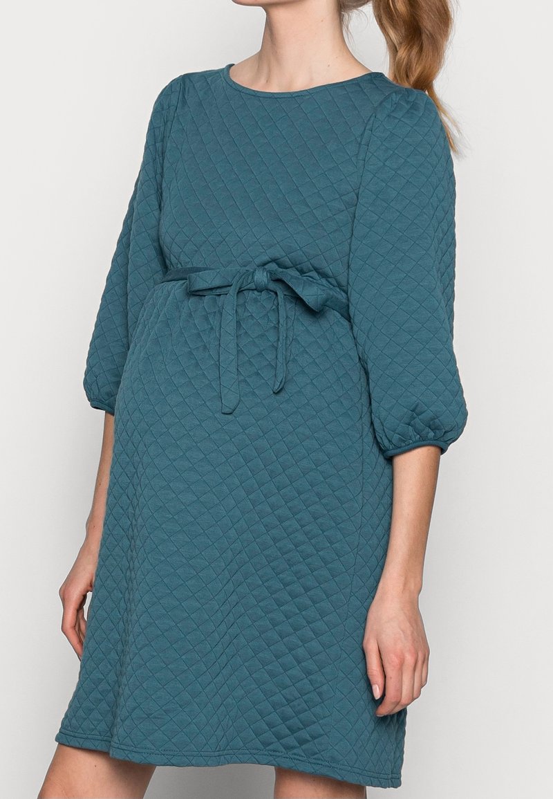 Robe matelassée bleu sarcelle avec manches trois-quarts, encolure ronde et détail de taille nouée. Texture douce et forme évasée.
