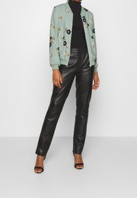 Hellgrüne Bomberjacke mit floralem Stickmuster, kombiniert mit schwarzen Lederhosen und schwarzen High Heels. Rollkragenpullover unter der Jacke.
