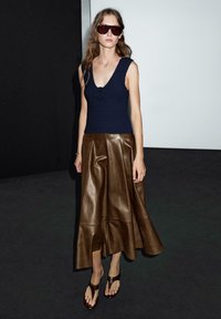 Námořnický pletený top s výstřihem do V a květinovým detailem, doplněný hnědou koženou midi sukní a tmavě hnědými plochými sandály. Doplňky zahrnují sluneční brýle.