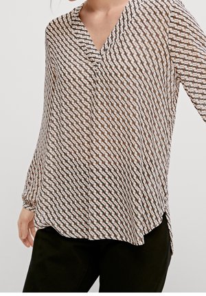 Kvinde iført bluse med lange ærmer med brun, sort og hvidt geometrisk mønster, kombineret med sorte bukser, poseret mod ensfarvet baggrund.