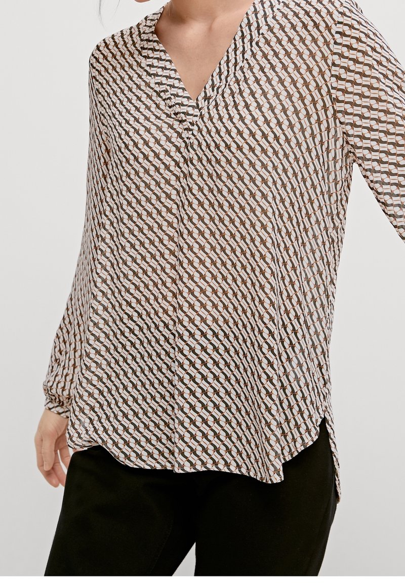 Femme portant un chemisier à manches longues avec un motif géométrique marron, noir et blanc, associé à un pantalon noir, posant devant un fond uni.