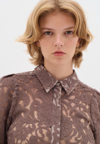 Blusa in pizzo di un morbido taupe con motivi floreali, colletto a polo, texture trasparente e vestibilità sagomata, con dettagli sottili sulle spalle.