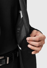 La mano di una persona che apre un blazer nero per rivelare l'etichetta interna e i dettagli del tessuto su uno sfondo semplice.