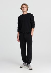 Felpa nera con colletto rotondo e polsini a costine, abbinata a pantaloni da jogging neri con vita elasticizzata e lacci, completata da sneakers grigie.