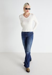 Balts adīkņu dzijas džemperis ar V-veida kakla izgriezumu, apvienots ar tumši ziliem platmalu džinsiem un brūniem potīšu zābakiem. Modelis valkā saulesbrilles.