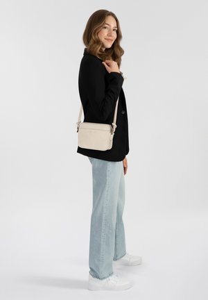 Beige schoudertas gemaakt van zacht materiaal, met een subtiel logo, verstelbare strap en rechthoekige vorm. Geshopt met een zwarte jas en lichte denim.