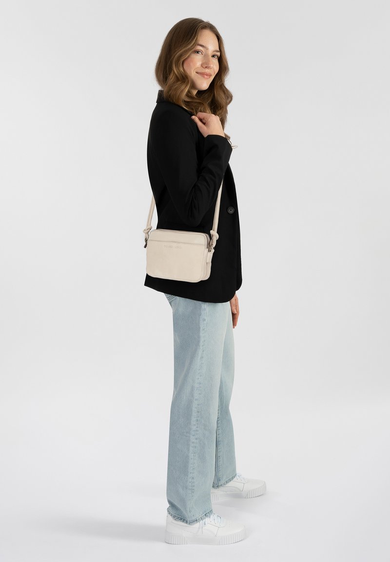 Borsa a tracolla beige realizzata in materiale morbido, con un logo discreto, tracolla regolabile e forma rettangolare. Indossata con una giacca nera e jeans chiari.