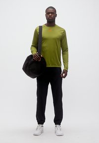 Άνδρας που φοράει πράσινο μπλουζάκι με μακριά μανίκια, μαύρο παντελόνι, λευκά αθλητικά παπούτσια, κρατώντας μια μεγάλη μαύρη τσάντα duffel στον ώμο, στέκεται σε λευκό φόντο.