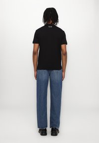 Tricou negru cu mânecă scurtă și un logo subtil pe spate, purtat cu blugi lejeri de culoare albastră deschisă și încălțăminte neagră.