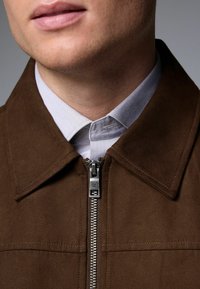 Brown suede jacket with a zip closure and pointed collar, layered over a light blue collared shirt. Notable textured fabric and hardware details.