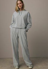 Svetlošedá skrátená mikina s kapucňou a šnúrkou, v kombinácii s voľnými šedými teplákmi. Hladká textúra látky, neformálny outfit, doplnený jemnými bočnými vreckami.