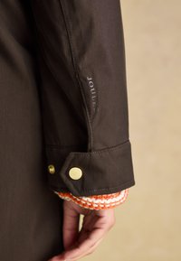 Manche de veste imperméable marron avec accents de quincaillerie dorée, logo "JOULES" brodé, avec un détail de poignet tricoté orange et blanc.