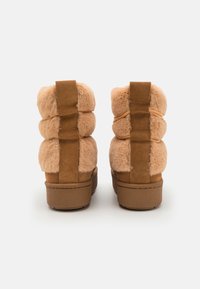 Bottines beiges avec une partie supérieure en fausse fourrure douce et des accents en daim. L'arrière présente des bandes élastiques et une semelle en caoutchouc robuste pour une meilleure adhérence.