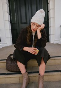 Rjava pletena torbica, sive široke hlače, kremasta rebrasta kapa s cofkom in taupe gležnjarji. V roki drži majhen kozarec kave.