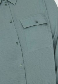 Shirt made of textured light teal fabric, featuring a button-down front, a single chest pocket, and round, matching buttons.