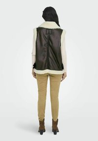 Bruine faux leren vest met een crèmekleurige schapenvacht kraag; de achterkant heeft een verticale naad en decoratieve strikaccenten aan de zijkanten.