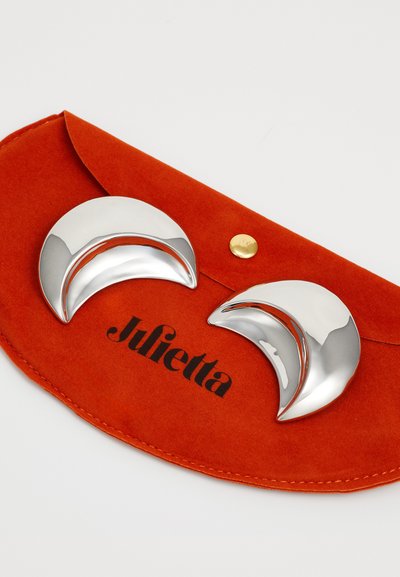 Boucles d'oreilles en forme de croissant argenté sur une pochette en suède orange avec un logo noir. Les boucles d'oreilles présentent des surfaces lisses et brillantes ainsi que des bords arrondis.