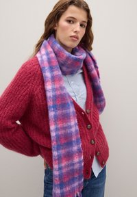 Giovane donna con capelli mossi, che indossa un cardigan rosso, una maglietta a righe blu, jeans blu e una sciarpa a quadri viola-rosa, in piedi contro un muro semplice.