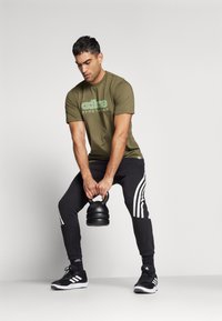 Maglietta sportiva verde oliva con logo "adidas sportear", pantaloni neri a sigaretta con strisce bianche e un kettlebell nero. Scarpe da ginnastica nere.