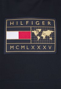 Marinblått tyg med en rektangulär patch där "HILFIGER" står i guld ovanför en röd och vit rand, med en världskarta och "MCMLXXXV" nedanför.