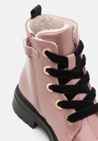 Bota de charol rosa con cordones de terciopelo negro, ojales dorados y forro de piel sintética. Presenta una cremallera lateral y una suela negra gruesa.