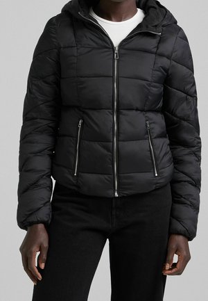 Persoon die een zwarte doorgestikte pufferjas met capuchon draagt, voorzien van een voorrits en ritssluitingen aan de zijzakken, over een wit shirt.