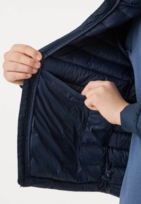 Mains tenant ouvert l'intérieur d'une veste matelassée bleu marine, montrant une poche intérieure et un cordon de serrage près de l'ourlet.