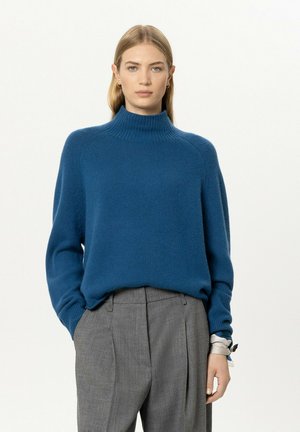 Woman with straight blonde hair wearing a blue turtleneck sweater and gray pleated trousers with hands in pockets against a plain background.
