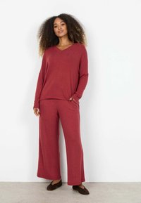 Kvinde med krøllet hår iført løs rød bluse med lange ærmer og bukser med vide ben, stående mod en hvid væg med den ene hånd i lommen.