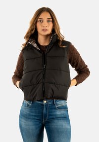 Gilet imbottito nero con chiusura a zip, dotato di una fodera a stampa leopardata, indossato sopra un maglione marrone a trama e abbinato a jeans blu.