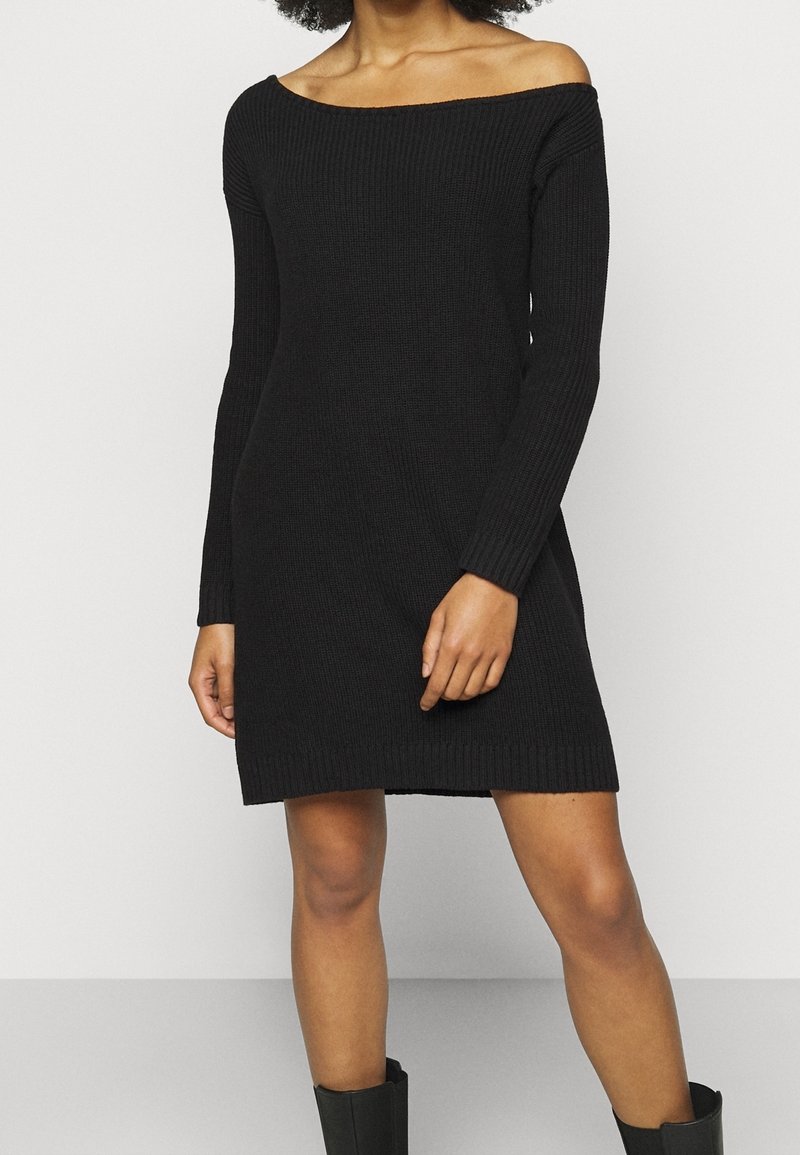 Femme portant une robe longue en maille noire à épaules dénudées et des bottes noires, se tenant devant un fond clair uni.