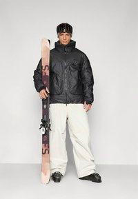 Čierna zateplená bunda typu puffer, biele snehové nohavice, držanie lyží s ružovo-čiernym dizajnom. Zahŕňa lyžiarske okuliare a čierne topánky.