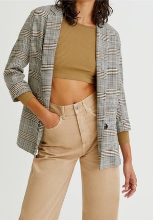 Woman wearing beige high-waist pants, cropped ribbed tan top, and three-quarter sleeve plaid blazer, with one hand in pocket and loose hair.