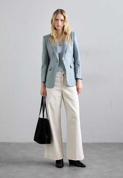 Blondynka w jasnoniebieskim żakiecie, prążkowej bluzce, szerokich beżowych spodniach, czarnych botkach, trzymająca czarną torebkę, stojąca na tle szarej podłogi i ściany.