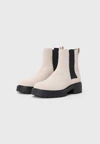 Beige semskede Chelsea-boots med sorte elastiske sidepaneler, tykke sorte gummisåler og trækstropper på en enkel lys baggrund.