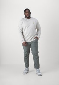 Felpa beige chiaro con un logo giallo sul petto, abbinata a pantaloni cargo verde oliva e sneakers bianche con accenti grigi.