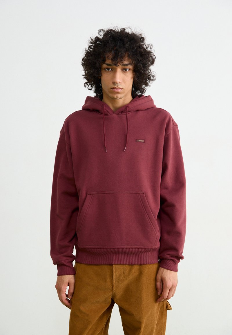 Burgundy hoodie made of cotton blend, featuring a drawstring hood, front pocket, and ribbed cuffs. Worn with beige corduroy pants.
