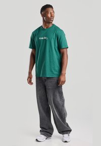 Green cotton t-shirt with white "Keith Haring" text, paired with loose-fitting dark gray jeans and white sneakers. Simple design, casual look.