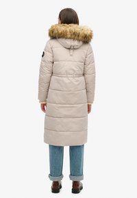Lang, beige puffjakke med en faux fur hætte. Har kviltet tekstur, metaltråde på hætten og en tætsiddende talje. Bæres med opfoldede jeans.