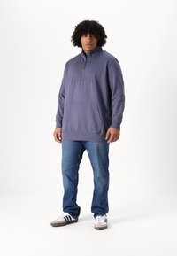 Sudadera gris con media cremallera en el cuello, con la palabra "ORIGINALS" en relieve en el frontal, combinada con jeans azules y zapatillas.