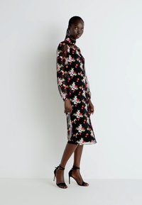 Vestido preto com estampado floral, mangas longas, transparência, bordado colorido e comprimento médio. Combinado com sandálias de salto alto pretas e com tiras.