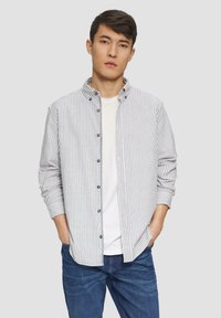 Camisa de rayas de manga larga en blanco y azul marino, con cuello alzado, cierre de botones y mangas remangadas sobre una camiseta blanca.