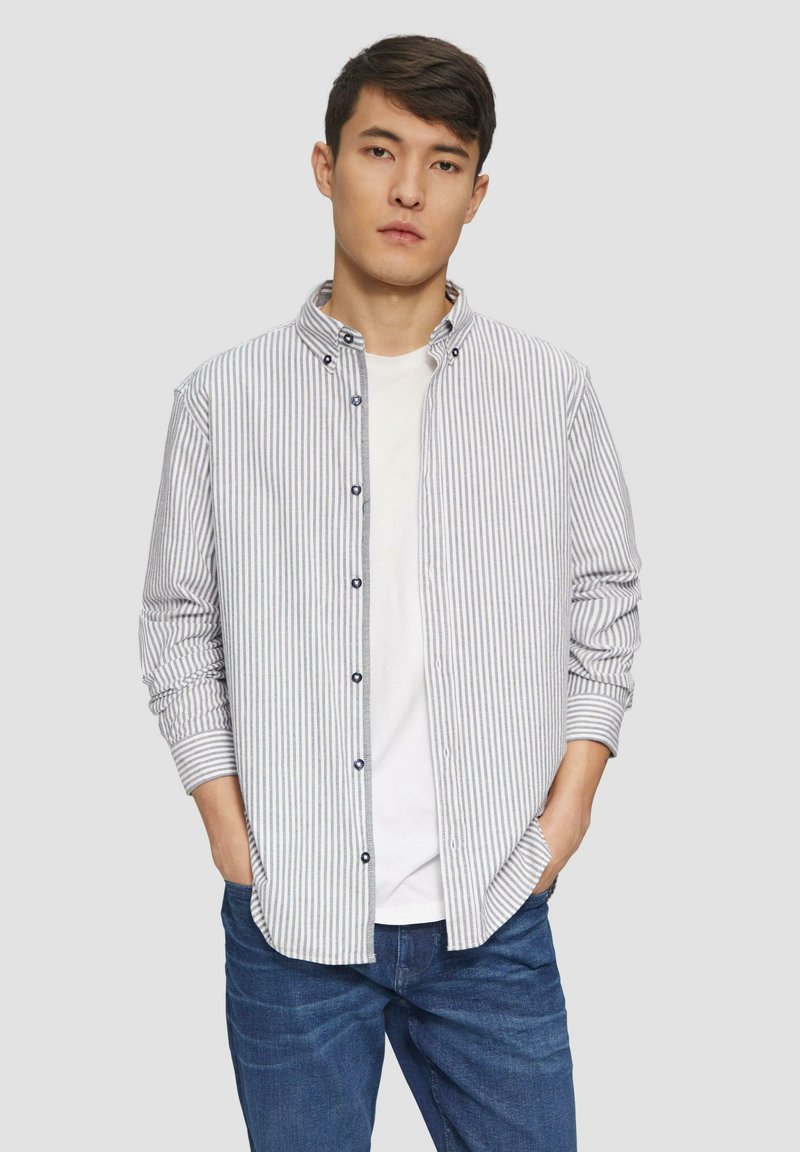 Camisa de rayas de manga larga en blanco y azul marino, con cuello alzado, cierre de botones y mangas remangadas sobre una camiseta blanca.